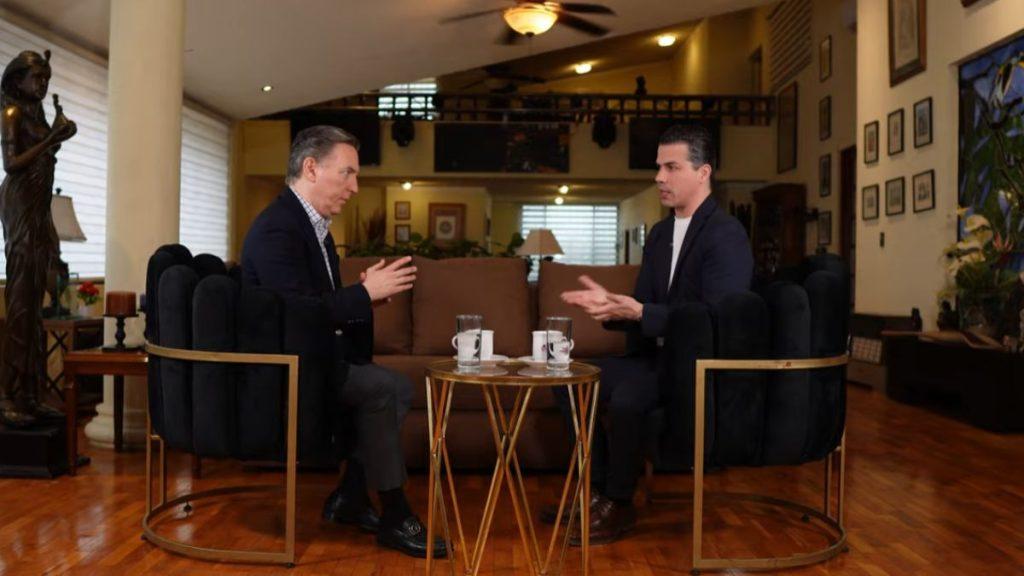 Two men in suits sit on dark velvet chairs facing each other in a spacious living room, clapping as they talk, with a small round table and mugs between them.