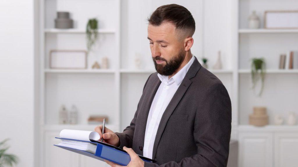 Bearded man in a gray suit signs documents in a bright office with shelves in the background.