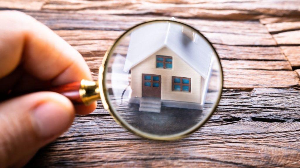 conoce-cuánto-vale-tu-casa-quantium-desarrollos-pedro-dávila - VOZ Y OPINION Hand holding a magnifying glass over a miniature house on a wooden surface, as if inspecting it closely.