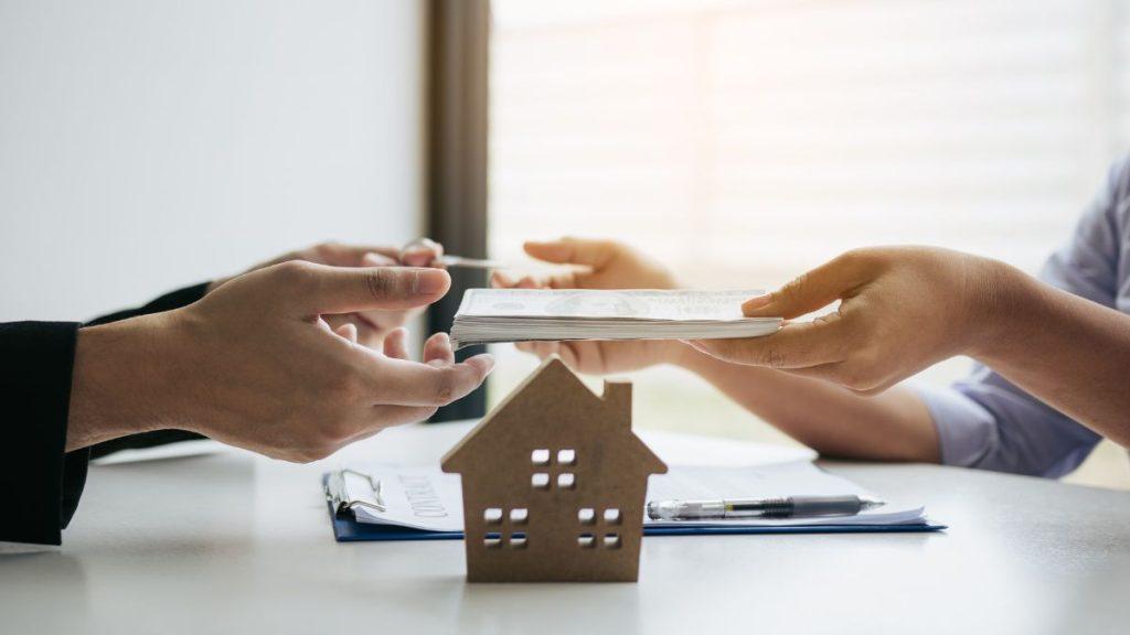 Two people exchange a stack of documents across a desk, with a small cardboard house model in the foreground—implies a real estate transaction.