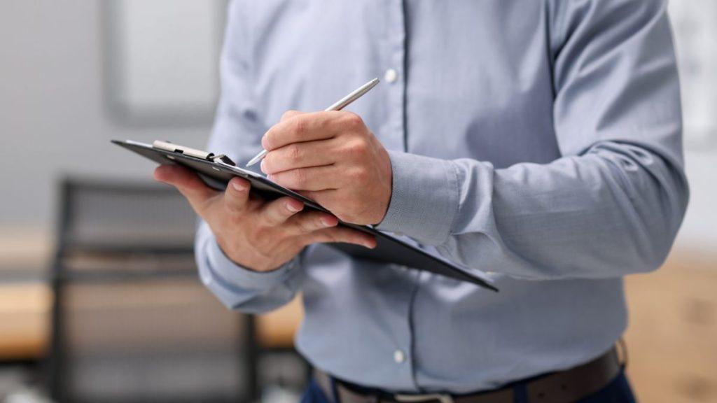 quanitum-desarrollos-avalúo-inmobiliario-contrato - VOZ Y OPINION Person in a light blue shirt writing on a clipboard in an office setting, taking notes with a pen held over the pad