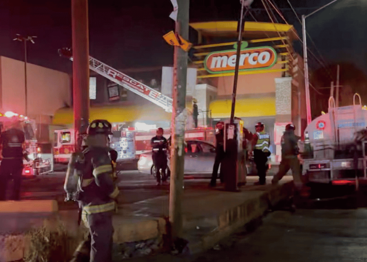 Nighttime emergency scene outside a Merco store with firefighters, police, and a fire truck responding to an incident.