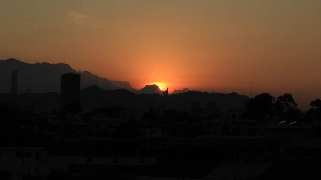 temperaturas-en-la-zona-metropolitana-de-monterrey 1 - VOZ Y OPINION Sunset over a city skyline with dark silhouettes of buildings and distant mountains, the orange sky fading to red near the horizon.